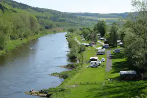 Campingplatz am Fluss mit Wohnmobilen und Stellplätzen auf der Wiese, Blick auf das Ufer und die grünen Hügel.