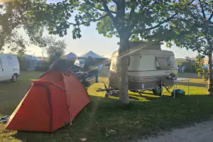 Rotes Zelt und Wohnwagen auf einem Campingplatz, daneben Auto und Campingstühle unter Bäumen.