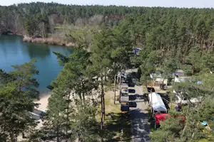 Campingplatz am See im Wald: Stellplätze mit Wohnwagen und Zelten, schmaler Weg und Bäume am Ufer.