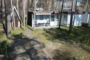 Wohnwagen mit überdachter Terrasse, zwei Stühlen und Tisch im Garten, umgeben von Bäumen und Zaun.