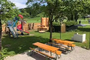 Campingplatz mit Rutsche, Spielbereich und Holztischen auf einer Wiese, umgeben von Bäumen und Hecken.