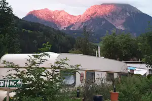 Weißer Wohnwagen auf dem Campingplatz mit Büschen und Blick auf bewaldete Berge im Abendlicht.