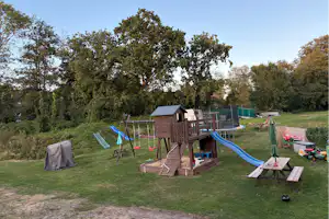 Spielplatz auf dem Campingplatz mit Holzspielturm, Rutschen, Schaukel und Wiese mit Zelten und Wohnwagen in der Nähe.