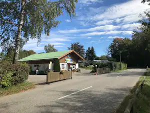 Eingang eines Campingplatzes mit kleinem Gebäude, Schildern und Einfahrt, umgeben von Bäumen und Hecken.