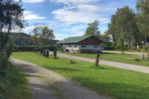 Campinghaus mit grünem Dach, Kiesweg und Rasenfläche, umgeben von Bäumen und Sträuchern auf dem Gelände.