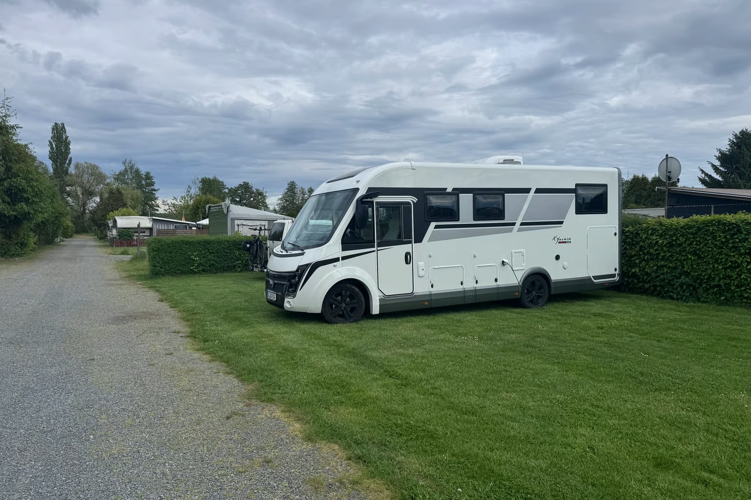 Weiße Wohnmobil-Stellfläche am Campingplatz, neben Hecke und Rasen, mit Kiesweg und weiteren Fahrzeugen im Hintergrund.