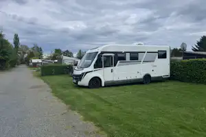Weiße Wohnmobil-Stellfläche am Campingplatz, neben Hecke und Rasen, mit Kiesweg und weiteren Fahrzeugen im Hintergrund.