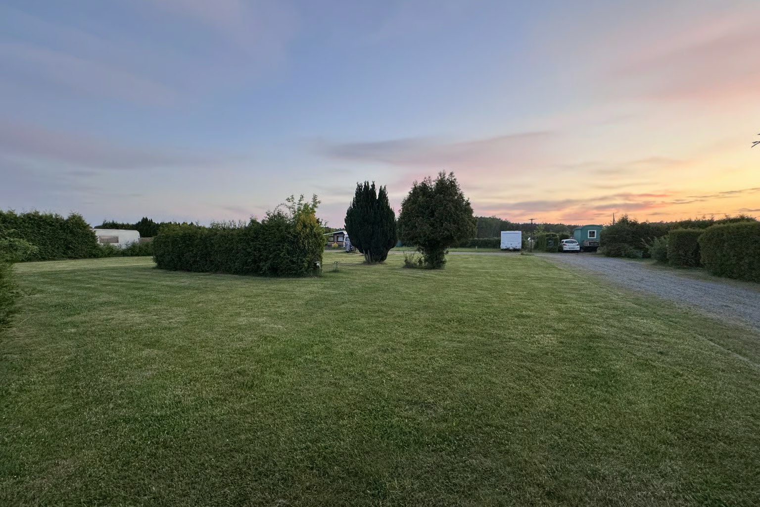 Grüner Campingplatz mit gepflegtem Rasen, Hecken und Bäumen, im Hintergrund Wohnwagen und ein Weg bei Sonnenuntergang.
