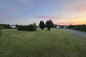 Grüner Campingplatz mit gepflegtem Rasen, Hecken und Bäumen, im Hintergrund Wohnwagen und ein Weg bei Sonnenuntergang.