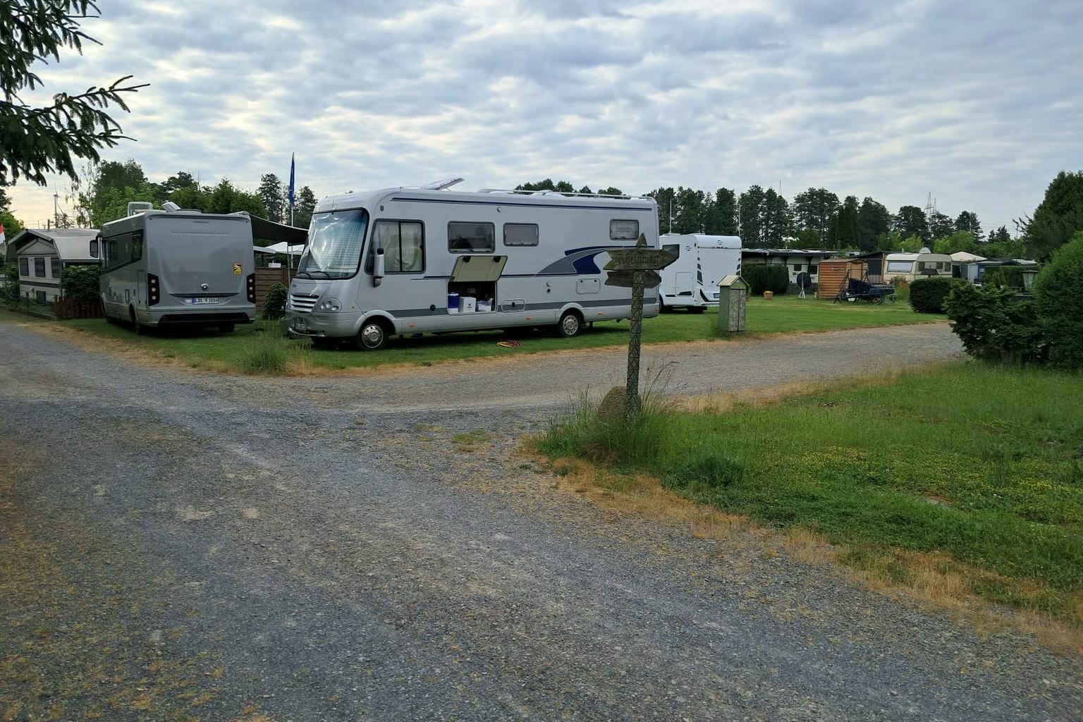 Wohnmobile und Campingbusse stehen auf einem Kiesplatz zwischen Rasenflächen, Bäumen und weiteren Stellplätzen.