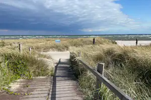 Holzweg durch Dünen zum Strand, dahinter das Meer und eine weite Himmelsszene mit Wolken.