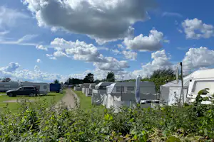 Campingplatz mit Wohnwagen und Wohnmobilen entlang eines Graswegs, im Hintergrund Bäume und große Wolken am Himmel.