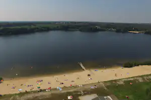 Sandstrand am See mit Badegästen, Steg und Blick über das Ufer mit Bäumen und Wiesen.