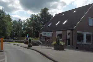 Campinggebäude mit Ziegeldach und Dachfenstern, Eingang und Schildern, davor gepflasterter Weg und Büsche im Grünen.