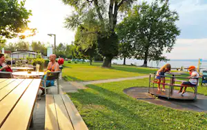 Camping-Terrasse mit Holztisch, Blick auf den Rasen und den See, im Hintergrund Bäume und Wohnwagen.