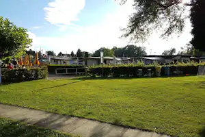 Grüner Rasen auf einem Campingplatz mit gepflastertem Weg, Hecken und mehreren Wohnwagen im Hintergrund.