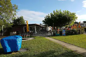 Terrasse am Campingplatz mit Sitzgarnituren, Sonnenschirm, Hecken und Rasenweg, im Hintergrund ein Gebäude und Bäume.