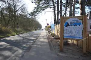 Schild „Camping Strandgut“ am Wegesrand, daneben Bäume und ein breiter Asphaltweg im Campingbereich.