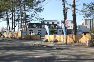 Campingplatz mit Wohngebäude im Hintergrund, Holzlattenzaun und Bäumen entlang des gepflasterten Wegs.
