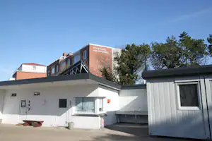 Campinggebäude mit Terrasse und Wohnwagen, weißes Haus mit Fenstern, daneben ein grauer Wohnwagen und Bäume im Hintergrund.
