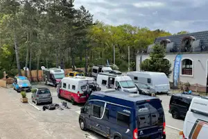 Reihe von Wohnmobilen und Transportern auf einem Campingplatz-Stellplatz, mit Bäumen im Hintergrund und Betonweg.