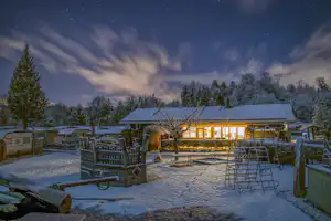 Winterlicher Campingplatz bei Nacht: beleuchtetes Restaurant, verschneite Wege und Zäune, Wohnwagen im Hintergrund und Sterne am Himmel.