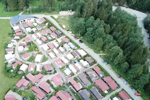 Luftaufnahme einer Campinganlage mit mehreren Gebäuden, roten Dächern, Zufahrtsstraße, Autos und Wald am Flussufer.