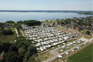 Luftaufnahme eines Campingplatzes mit vielen Stellplätzen, Wegen und Wohnwagen nahe einem breiten Fluss und einem Ort am Ufer.