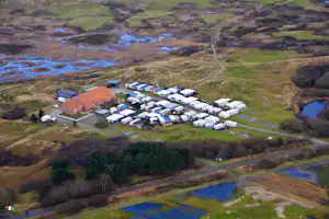 Luftaufnahme eines Campingplatzes mit Wohnwagen, Stellplätzen, Wegen und einem Gebäude inmitten von Wiesen und Bäumen.