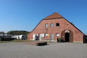 Ziegelgebäude mit großem Tor und Fenstern, davor gepflasterter Platz, Mülltonnen und Absperrzaun am Campingplatz.