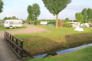 Campingplatz mit Wohnwagen und Zelt auf dem Stellplatz, Rasenfläche, Bäume und ein kleiner Teich neben dem Holzsteg.
