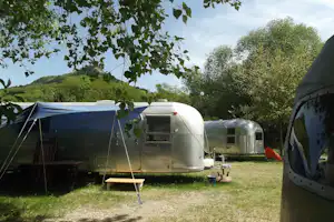 Silberner Airstream und Zelt mit Vorzelt auf dem Campingplatz, umgeben von Bäumen und Blick auf grüne Hügel.