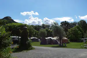 Zelt auf dem Campingplatz zwischen Bäumen, daneben Wohnwagen und Autos, im Hintergrund Wiese und große Wolken am Himmel.