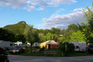 Campingplatz mit Zelt und Wohnwagen, Autos auf dem Stellplatz, Bäume und ein bewaldeter Hügel unter blauem Himmel.