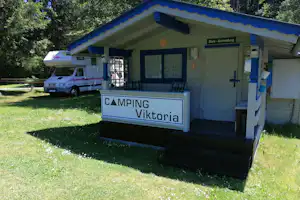 Camping-Viktoria Wohnwagen mit Vorzelt und Schild am Eingang, davor Rasen und ein Wohnmobil auf dem Stellplatz im Grünen.