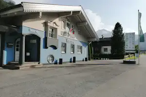 Campinggebäude mit Eingangsvorbau, Flaggen am Haus und breitem Parkplatz vor dem Gebäude, im Hintergrund Bäume und Berge.