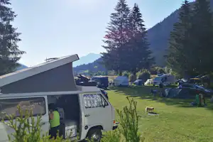 Wohnmobil auf einem Campingplatz mit grünem Rasen, weiteren Fahrzeugen und Zelten, umgeben von hohen Tannen und Bergen.