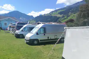 Mehrere Wohnmobile auf einer Campingwiese, im Hintergrund ein Gebäude mit Schild und grüne Berge.