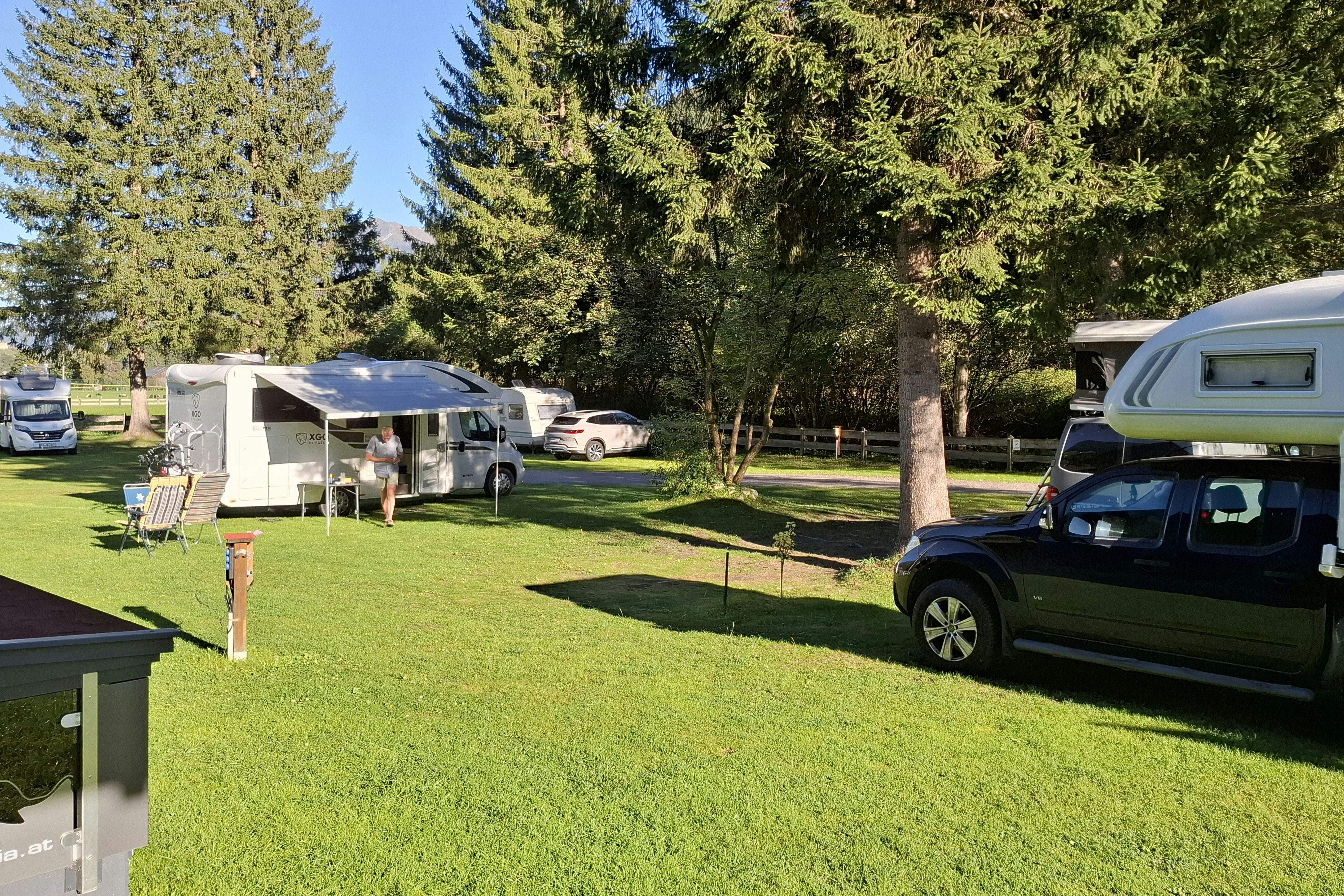 Wohnwagen und Zelte auf einem grünen Campingplatz, daneben ein grüner Camper-Van und Bäume im Hintergrund.