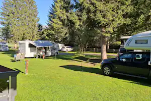 Wohnwagen und Zelte auf einem grünen Campingplatz, daneben ein grüner Camper-Van und Bäume im Hintergrund.