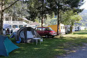 Campingzelt und weitere Wohnwagen auf einem Stellplatz im Grünen, mit Bäumen, Autos und einem weißen Gebäude im Hintergrund.