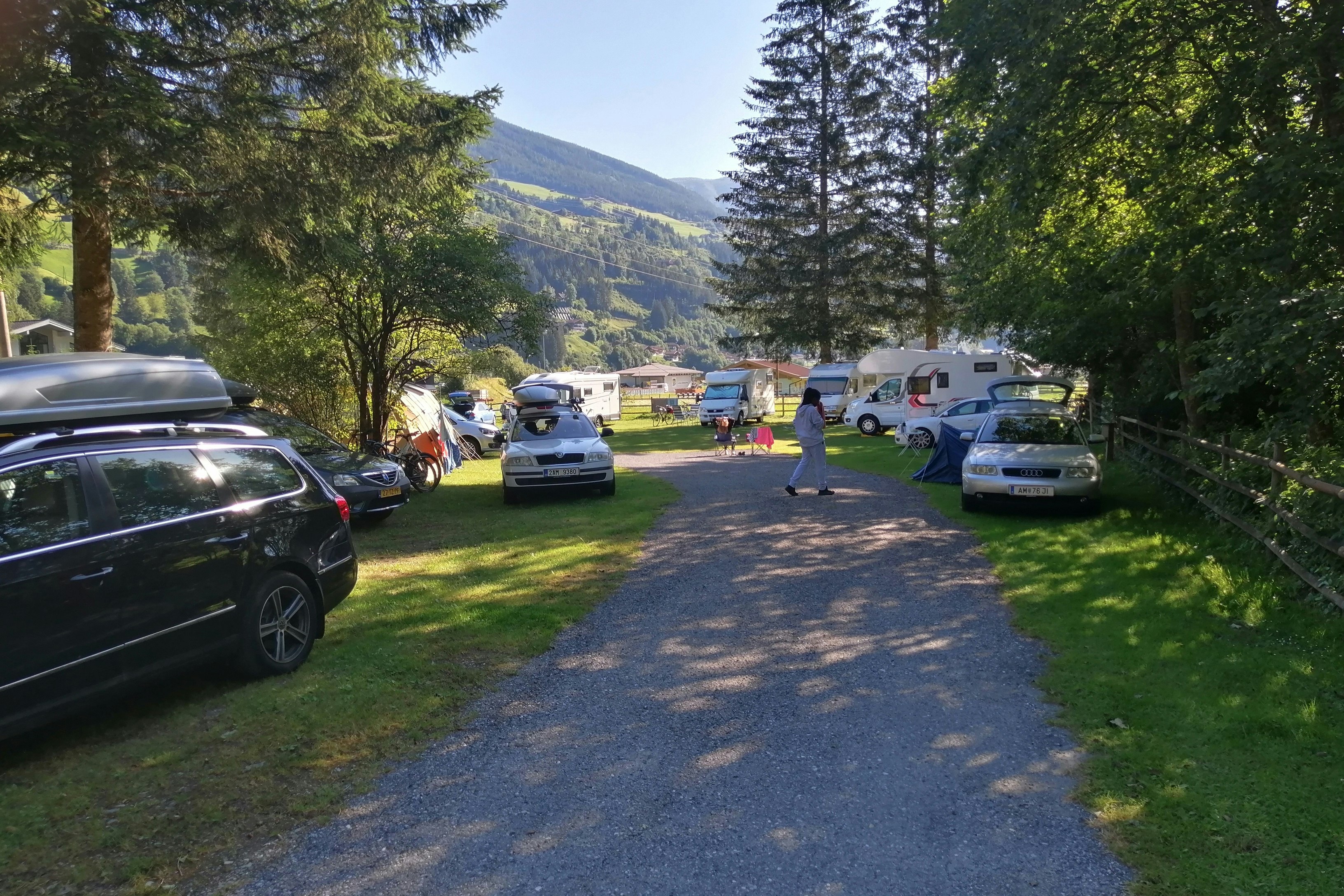 Campingplatz-Zufahrt mit Autos und Wohnmobilen, Stellplätzen am Weg, Bäume und Berge im Hintergrund.