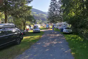 Campingplatz-Zufahrt mit Autos und Wohnmobilen, Stellplätzen am Weg, Bäume und Berge im Hintergrund.