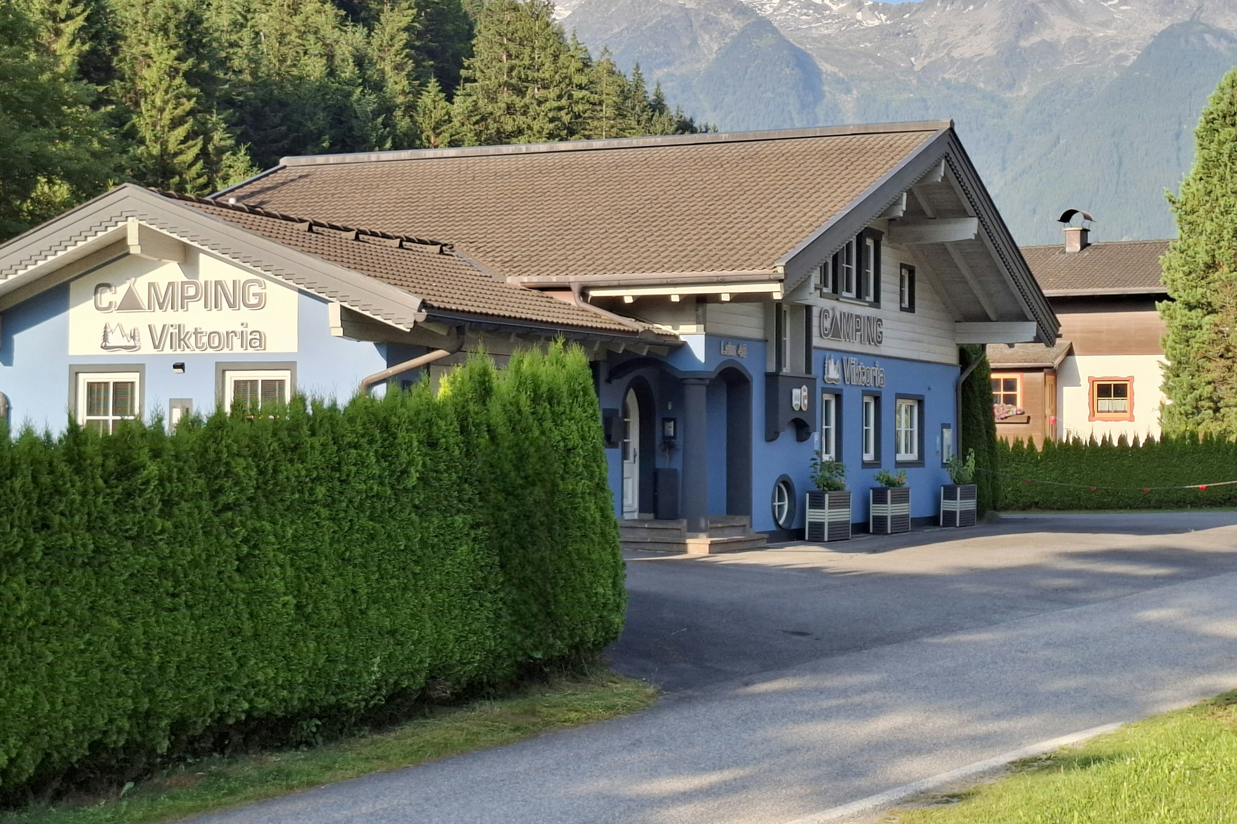 Campinggebäude mit Schild „Camping La Vignette“ und Eingang, davor Hecke und Auffahrt, im Hintergrund Berge und Wald.