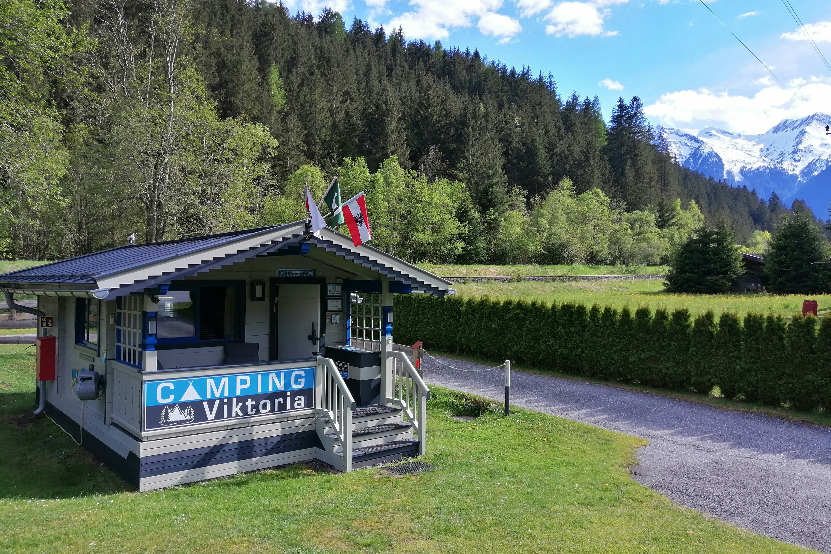 Camping-Viktoria Mobilheim mit Veranda und Stufen, Rasenfläche und Kiesweg, umgeben von Hecken, Bäumen und Bergen im Hintergrund.