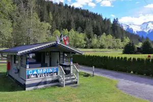Camping-Viktoria Mobilheim mit Veranda und Stufen, Rasenfläche und Kiesweg, umgeben von Hecken, Bäumen und Bergen im Hintergrund.