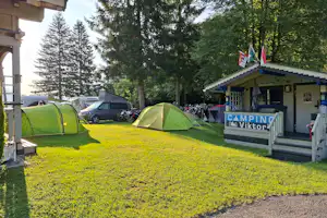 Campingplatz mit zwei grünen Zelten, Auto und Rezeption mit Schild „Camping“ am Rand, umgeben von Bäumen.