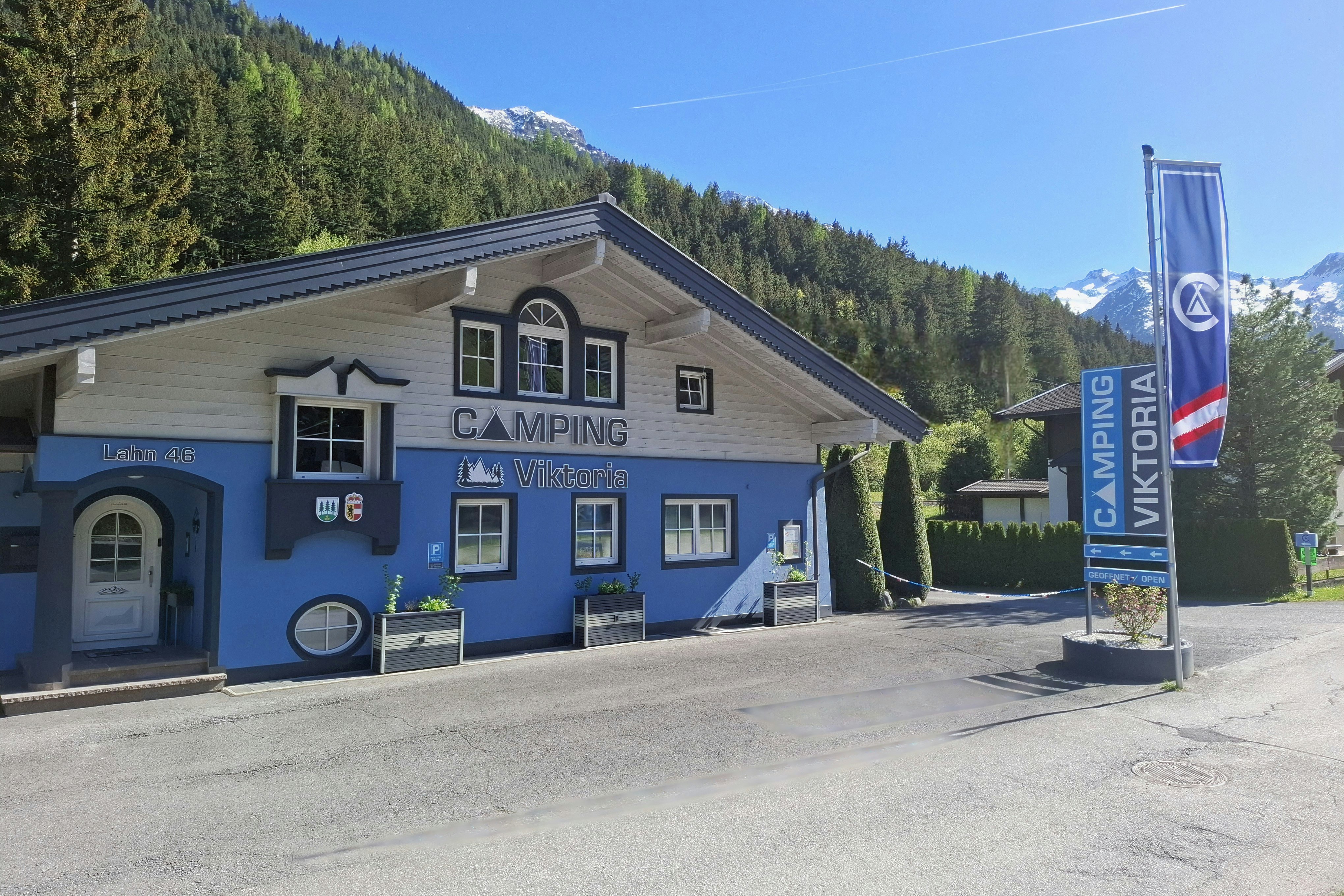 Blaues Campinggebäude mit Schildern „Camping“ und „Willkommen“, davor Betonplatz, Berge und Nadelwald im Hintergrund.