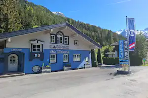 Blaues Campinggebäude mit Schildern „Camping“ und „Willkommen“, davor Betonplatz, Berge und Nadelwald im Hintergrund.