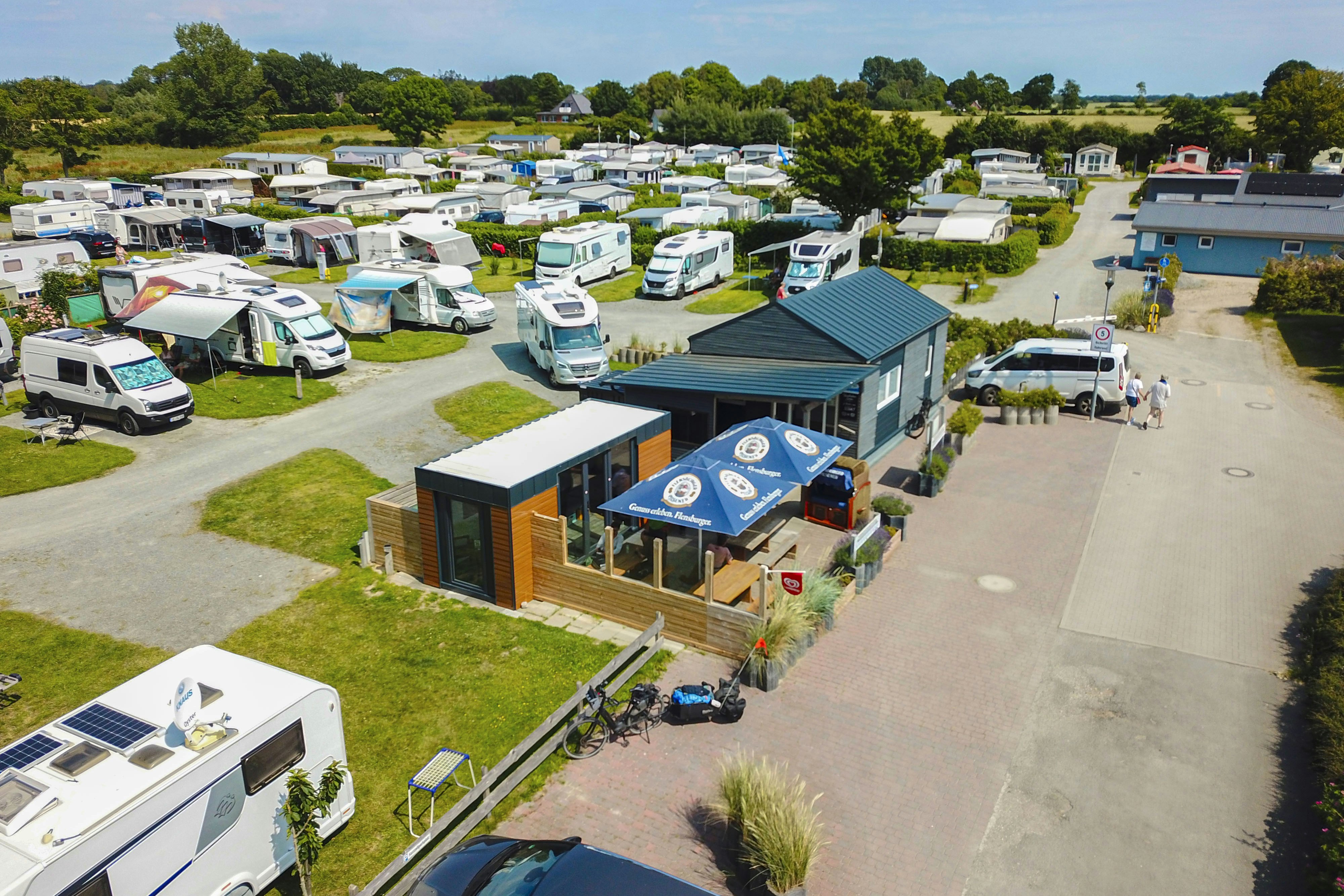 Campingplatz mit Wohnmobilen und Stellplätzen, Rezeption mit Glasfront und Sonnenschirmen, gepflasterte Wege und Rasenflächen zwischen Bäumen.